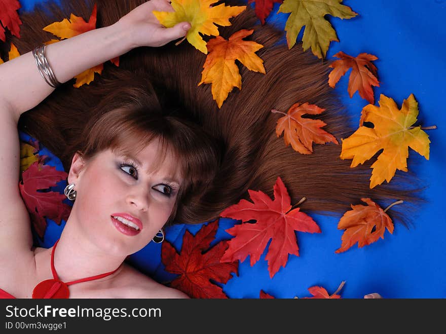 Cute brunette girl with hair spread around, laying on blue background, surrounded by maple leaves. Cute brunette girl with hair spread around, laying on blue background, surrounded by maple leaves