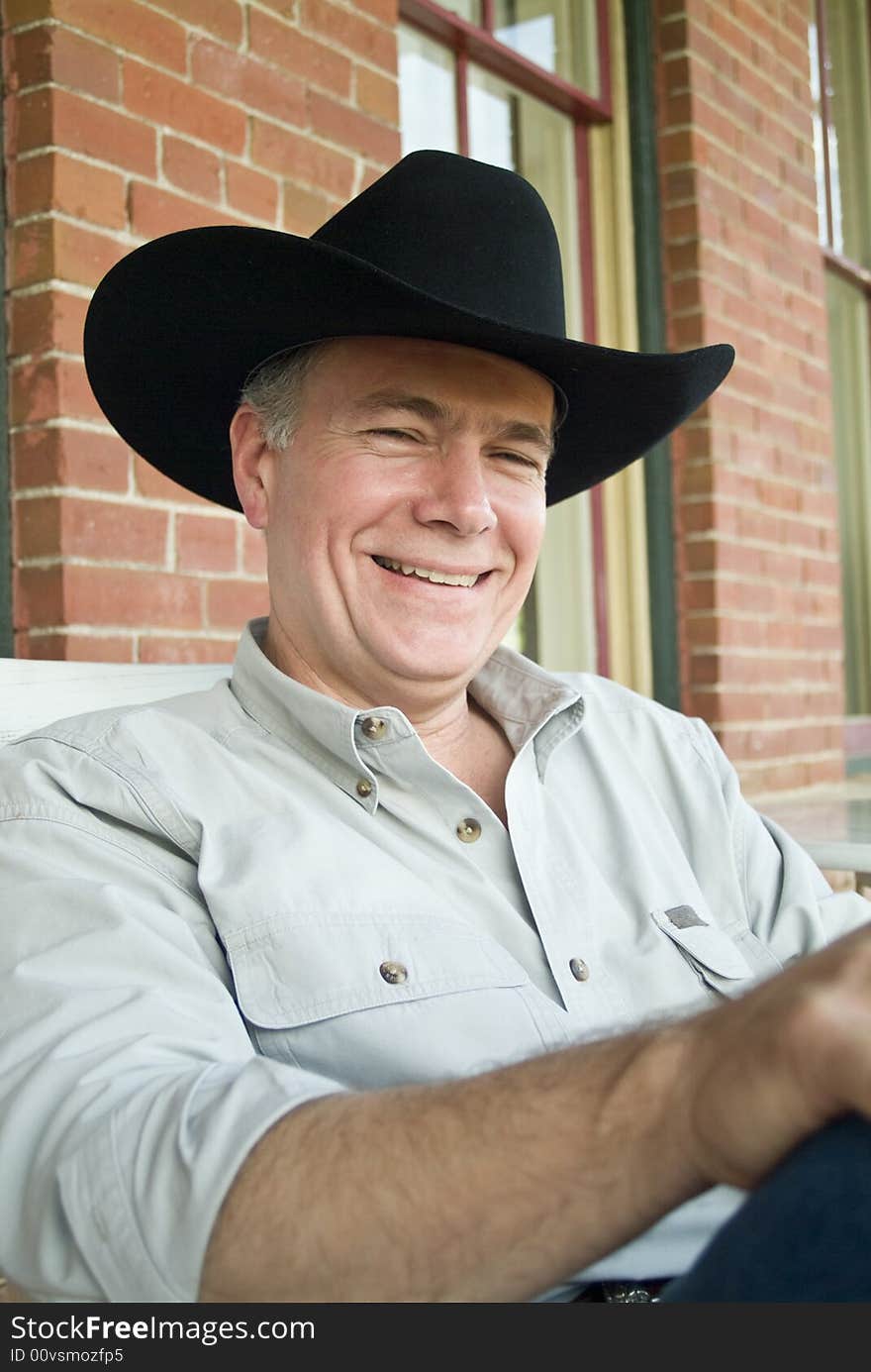 A man wearing a cowboy hat, sitting on a porch, with gentle laughter covering his face. A man wearing a cowboy hat, sitting on a porch, with gentle laughter covering his face.