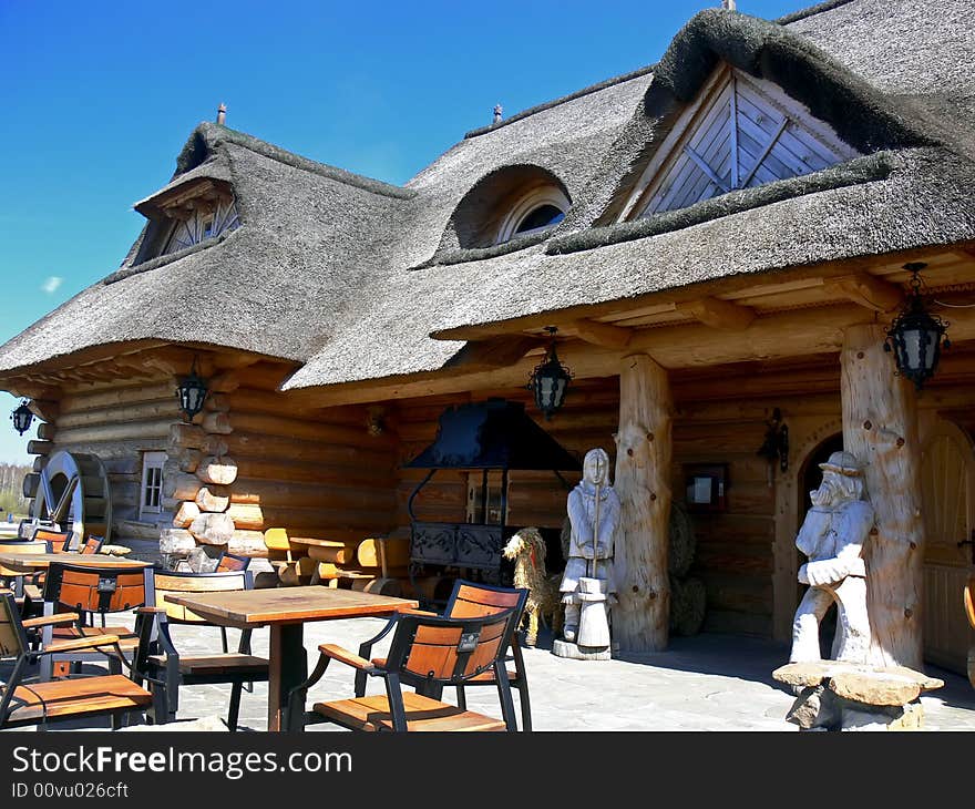 The old wooden house with mill wheel. Terrace in front of the building with chairs and tables. Two figures carved from wood.