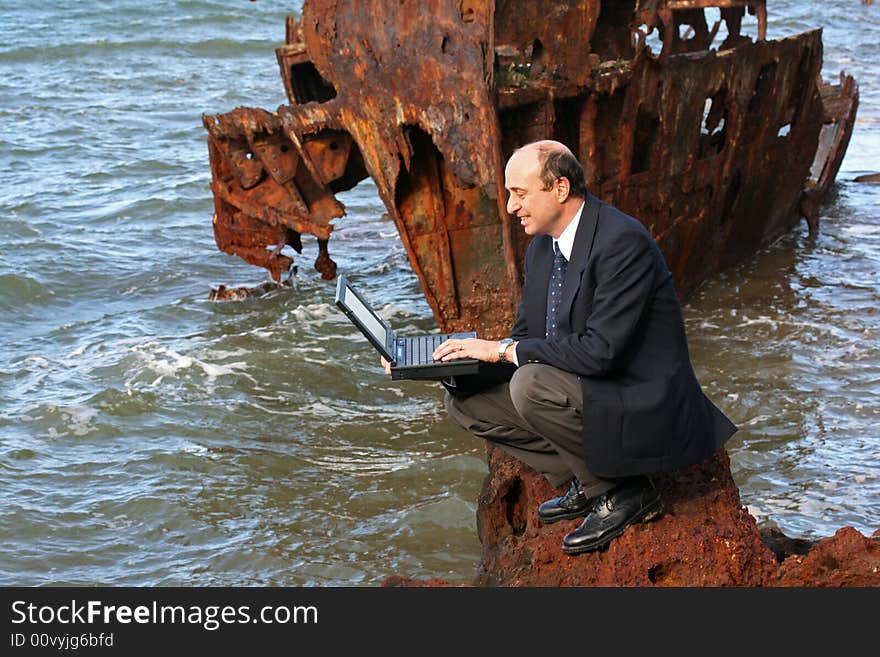 Businessman sitting on the rocks at the beach with his laptop and with a beautiful old rusting shipwreck in the background. Businessman sitting on the rocks at the beach with his laptop and with a beautiful old rusting shipwreck in the background