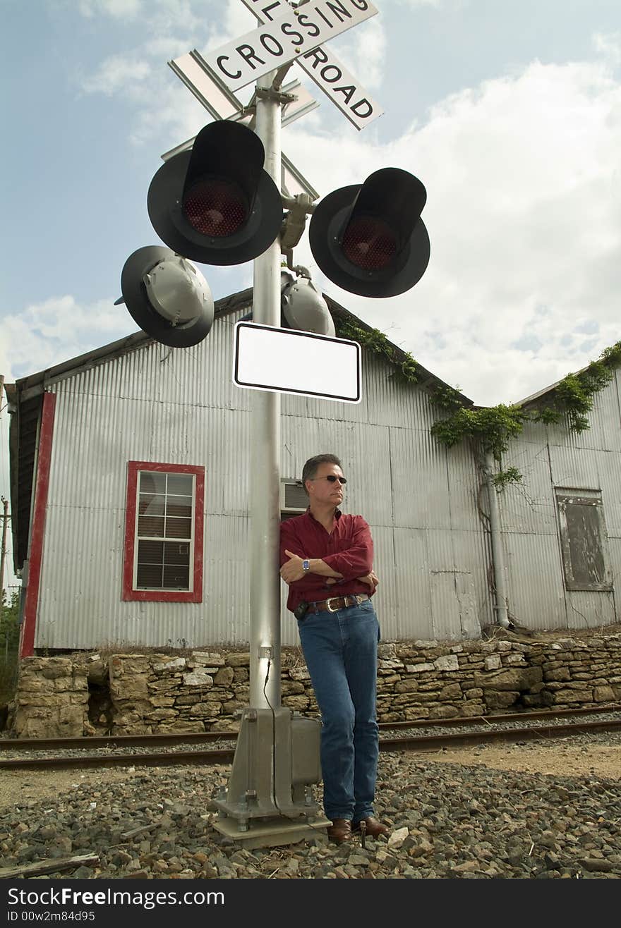 A man wearing dark sunglasses, leaning up against a railroad crossing signal lights pole.