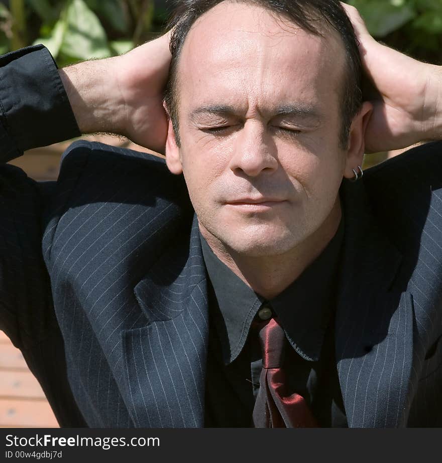 A shot of a business man taking in the Sun outdoors.  His hand are resting on the back of his head and his eyes are closed.