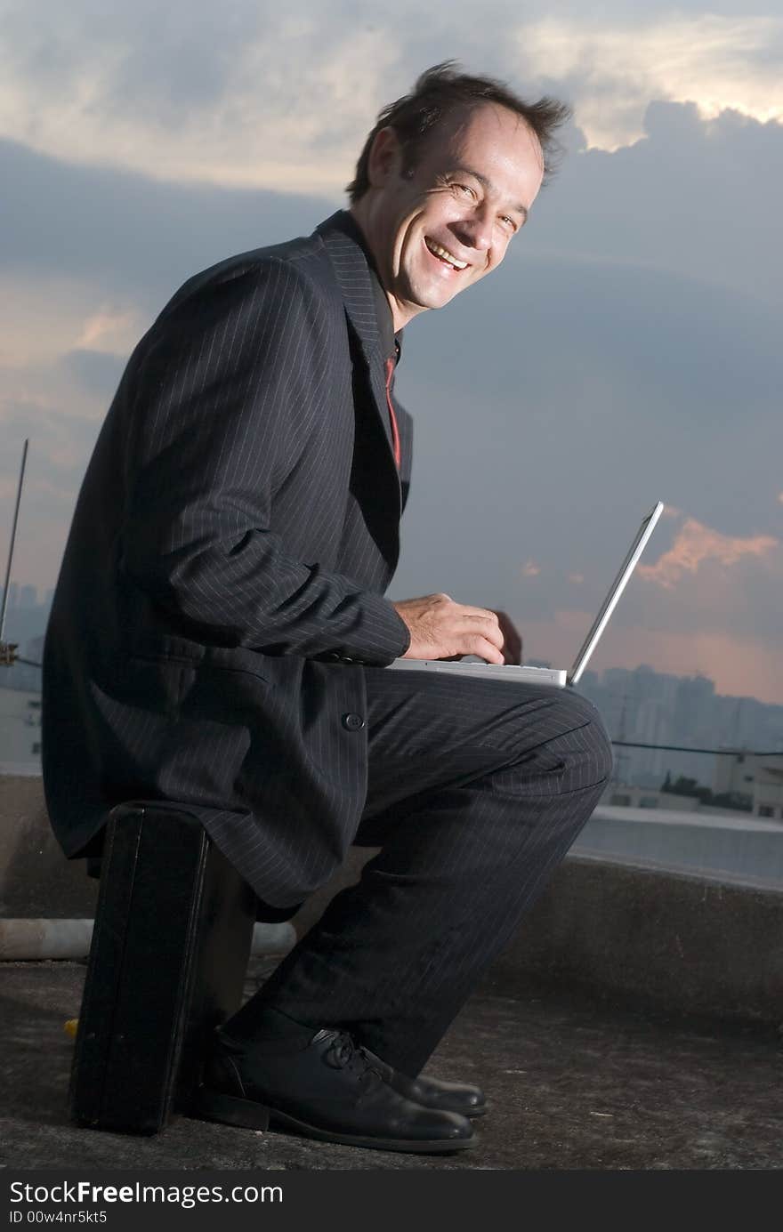 A business man, sitting on his briefcase, working on his laptop outdoors, while smiling. A business man, sitting on his briefcase, working on his laptop outdoors, while smiling.