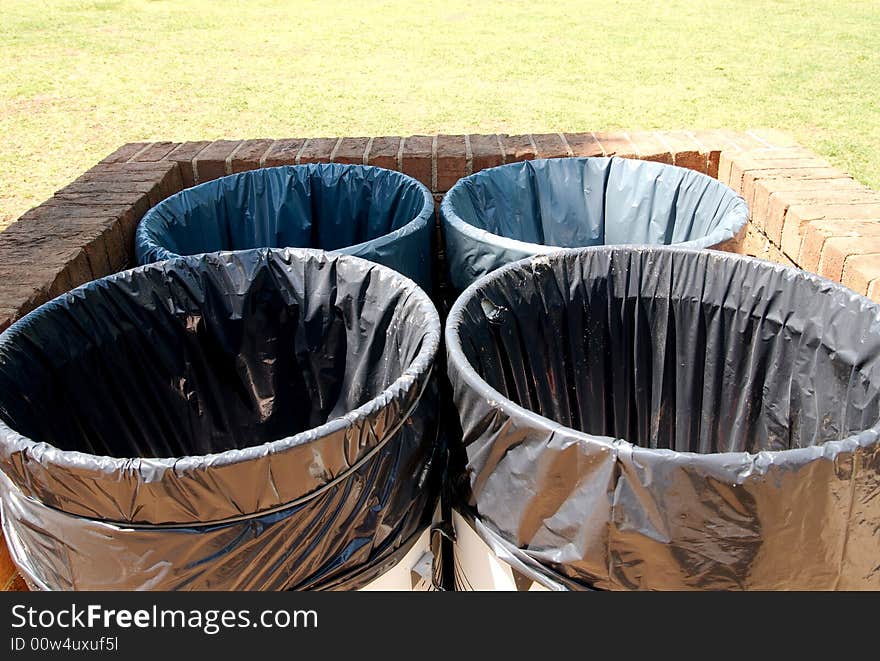 Four trash cans together at a park in the summer.