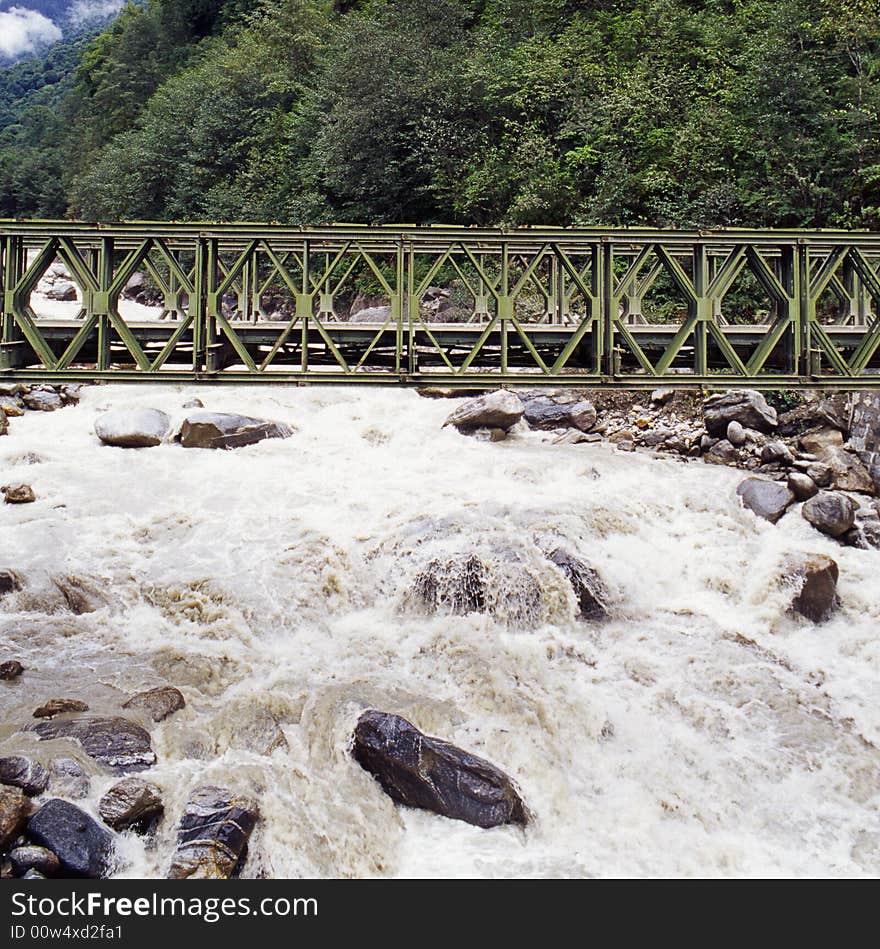 It is a metal bridge on the river, it is in Tibet of China. with green forest.