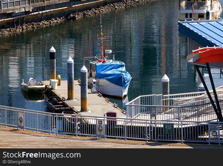 Boats Moored at Pier in Seattle show a couple of small boats tied up at the docks with many beautiful reflections showing across the water in this beautiful waterfront composition. A fence frames the immediate foreground.