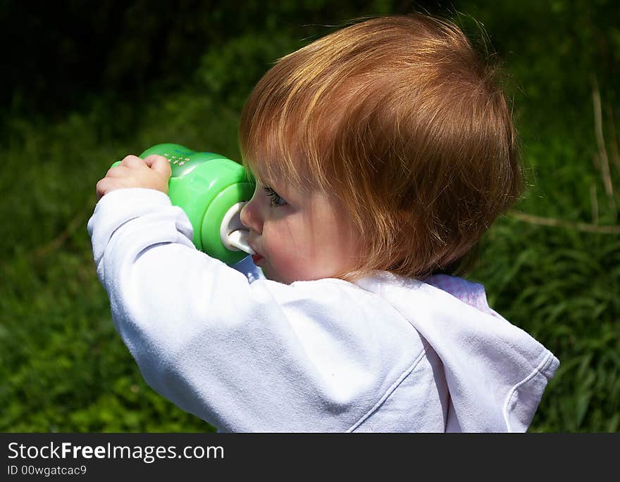 The little girl drinks from a green bottle on a background of a grass