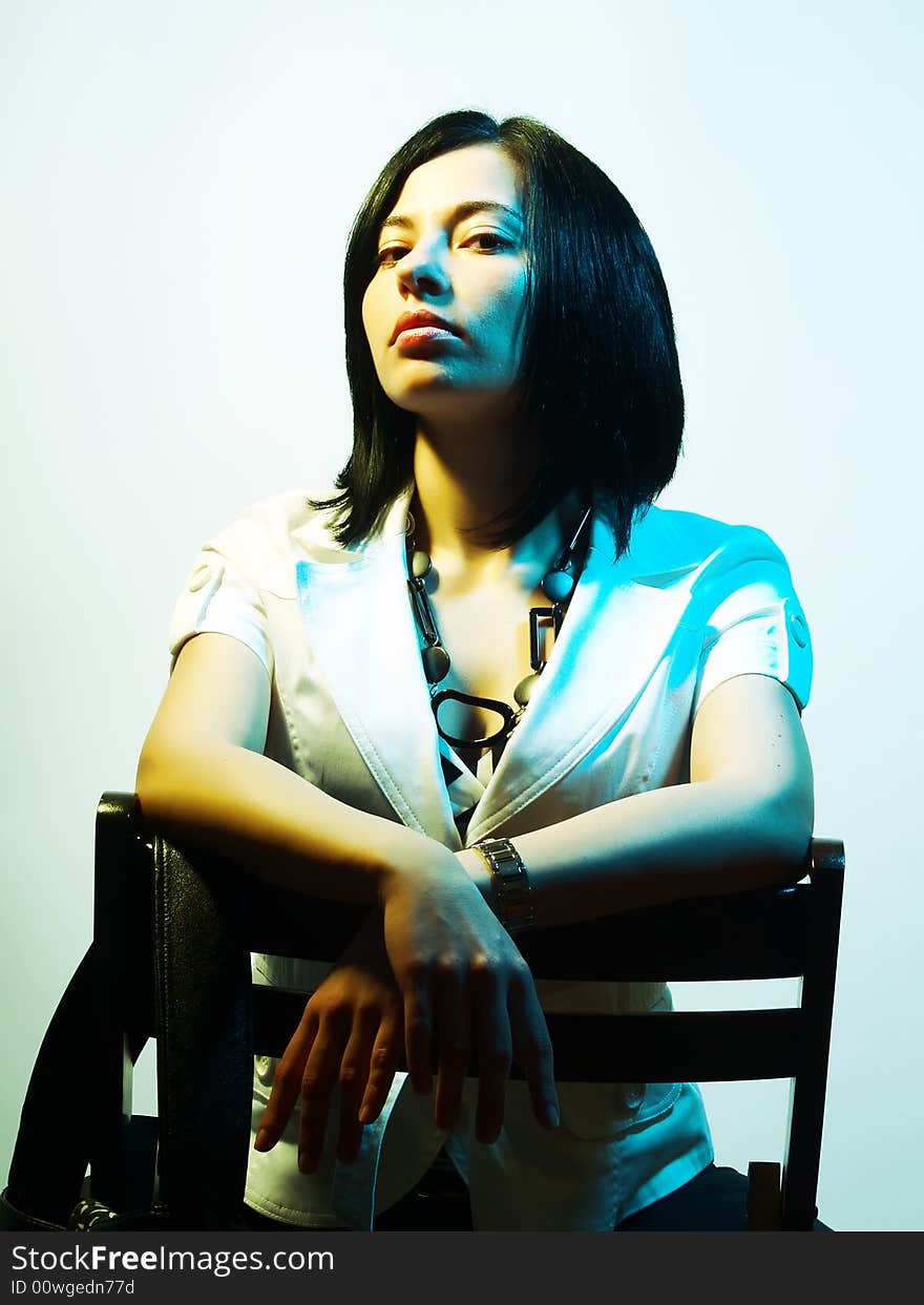 A high-key portrait about an attractive trendy girl with black hair who is lighted with blue and yellow, she is sitting on a chair and she has a charming look. She is wearing black pants, a white coat and a stylish necklace. A high-key portrait about an attractive trendy girl with black hair who is lighted with blue and yellow, she is sitting on a chair and she has a charming look. She is wearing black pants, a white coat and a stylish necklace.