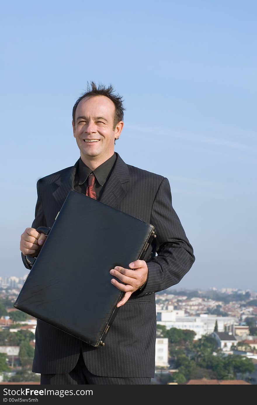 Businessman standing on a city rooftop and smiling while holding his briefcase close to his chest. Businessman standing on a city rooftop and smiling while holding his briefcase close to his chest