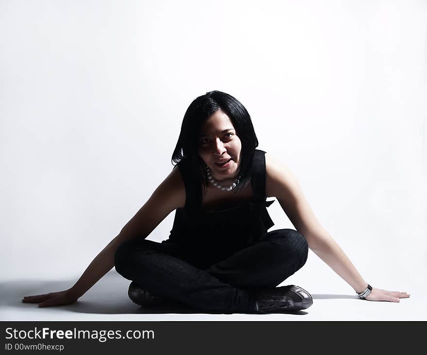 A portrait about an attractive trendy girl with black hair who is sitting, she is spreading her arms on ground and she is smiling. She is wearing a black dress, blue jeans and a white necklace. A portrait about an attractive trendy girl with black hair who is sitting, she is spreading her arms on ground and she is smiling. She is wearing a black dress, blue jeans and a white necklace.