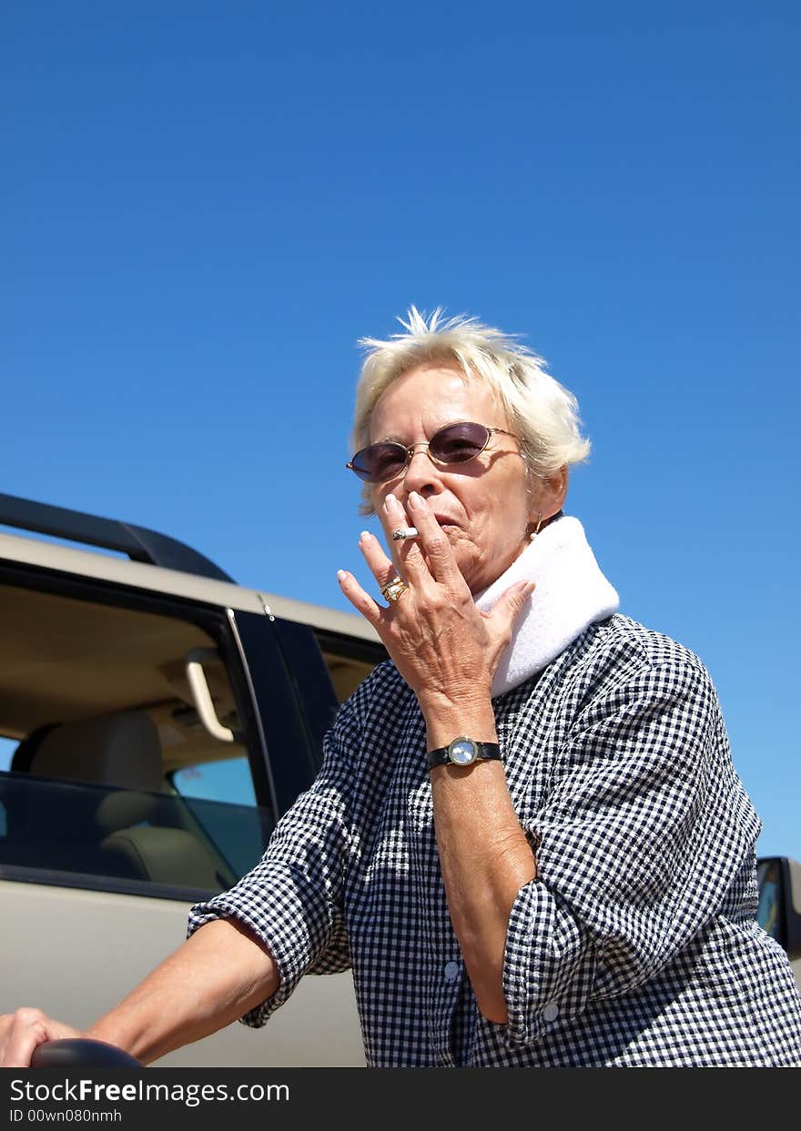 Older woman with close-cropped white hair taking a drag from a cigarette while standing outside in the desert. Shot is framed against a clear blue sky and desert sand. Older woman with close-cropped white hair taking a drag from a cigarette while standing outside in the desert. Shot is framed against a clear blue sky and desert sand.