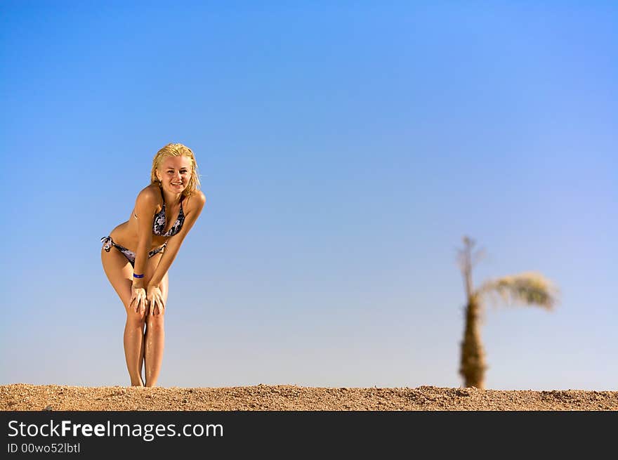 Young woman standing on sand leaning on her knees and waiting for something. Blue sky and palm on a background. there is a good place for your text