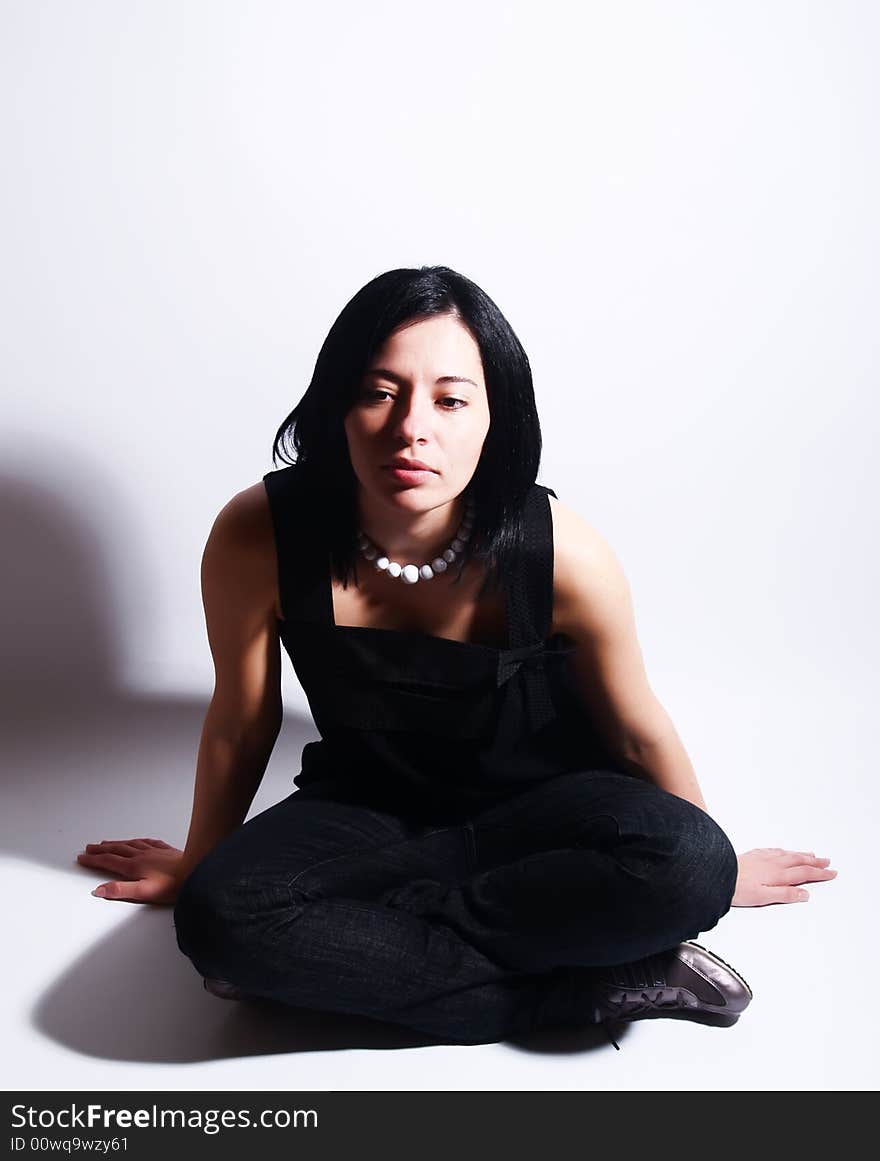 A high-key portrait about an attractive trendy girl with black hair who is sitting, she is spreading her arms on ground and she is thinking. She is wearing a black dress, blue jeans and a white necklace. A high-key portrait about an attractive trendy girl with black hair who is sitting, she is spreading her arms on ground and she is thinking. She is wearing a black dress, blue jeans and a white necklace.