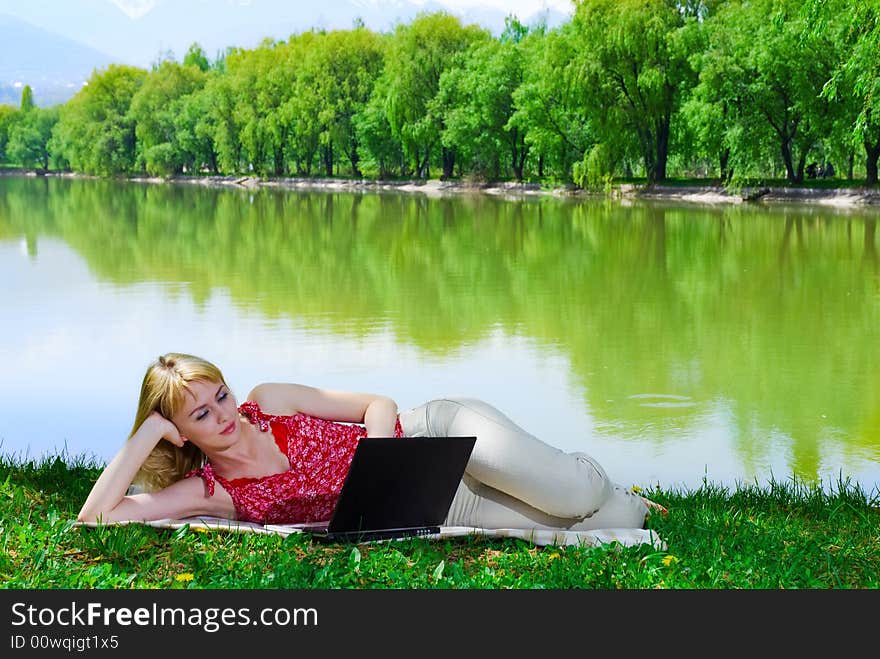 Beautiful young woman with laptop near the lake laying on the grass