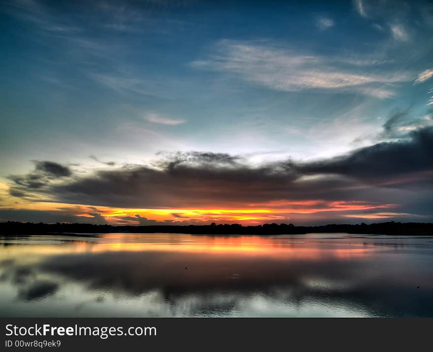 Sun setting below low altitude clouds while illuminating high altitude ones over a still lake. Sun setting below low altitude clouds while illuminating high altitude ones over a still lake