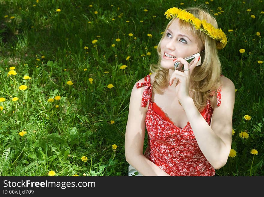 Young beautiful blond hair woman in garland talking on the mobile phone, sitting on the grass among dandelions. Young beautiful blond hair woman in garland talking on the mobile phone, sitting on the grass among dandelions