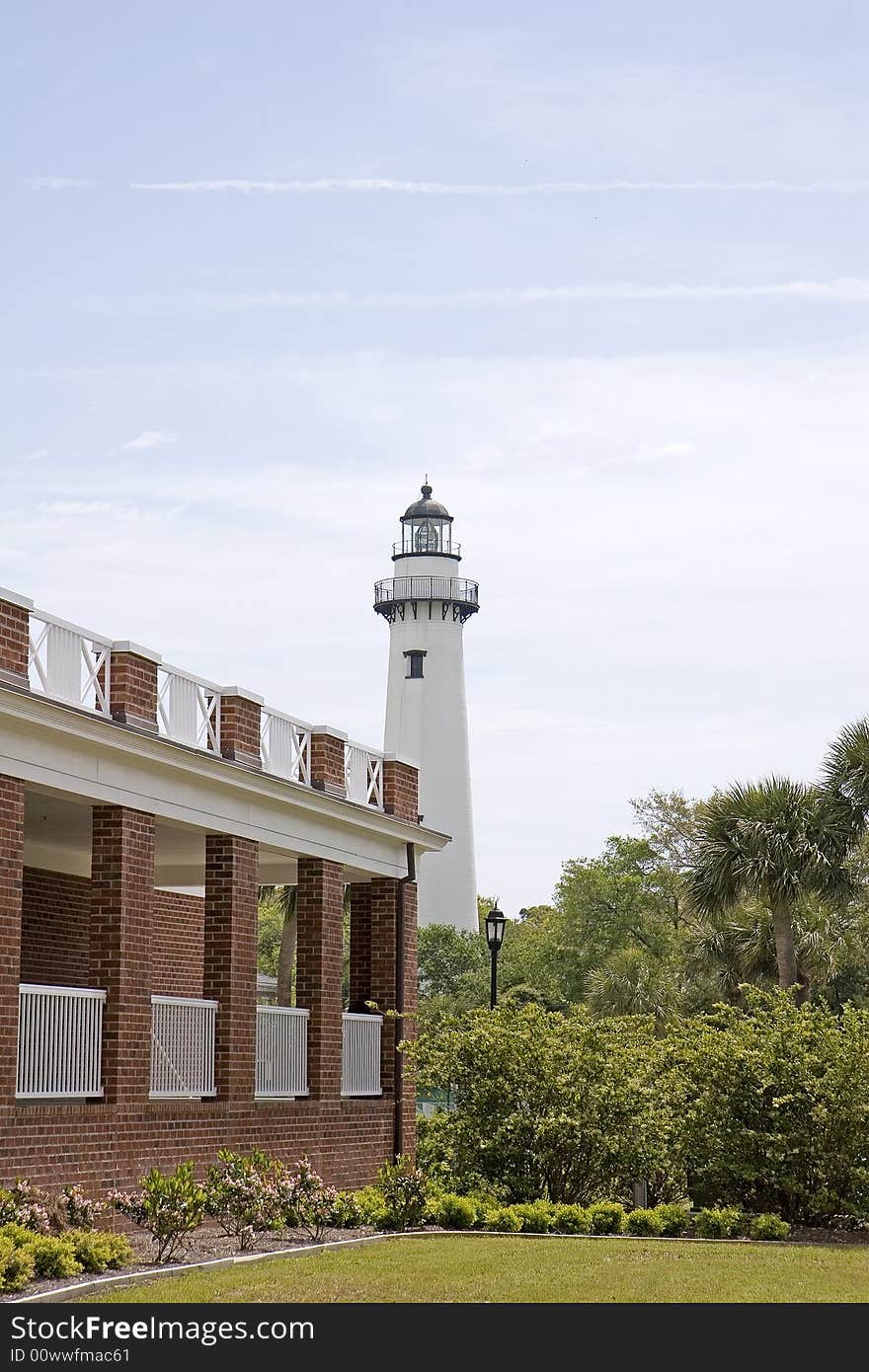 A lighthouse on the horizon past a brick building and trees. A lighthouse on the horizon past a brick building and trees