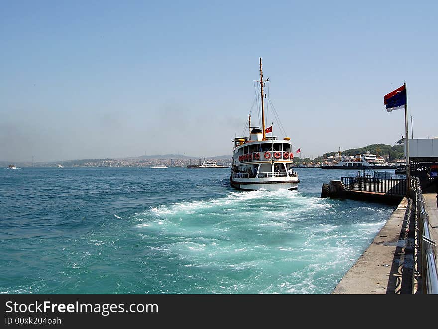 Commuter boat departing from the pier with blue and red flags, boat's propeller churn the blue sea leaving a green trail and white waves behind, in Eminonu, Istanbul. Commuter boat departing from the pier with blue and red flags, boat's propeller churn the blue sea leaving a green trail and white waves behind, in Eminonu, Istanbul