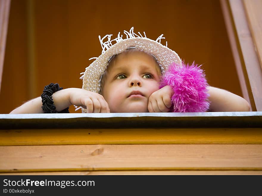 The little girl in a summer hat looks out of a window of a wooden country house. The little girl in a summer hat looks out of a window of a wooden country house