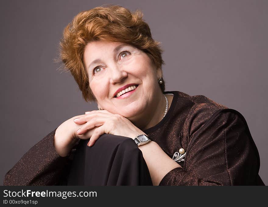 The cheerful elderly woman in a brown dress on a grey background. The cheerful elderly woman in a brown dress on a grey background.