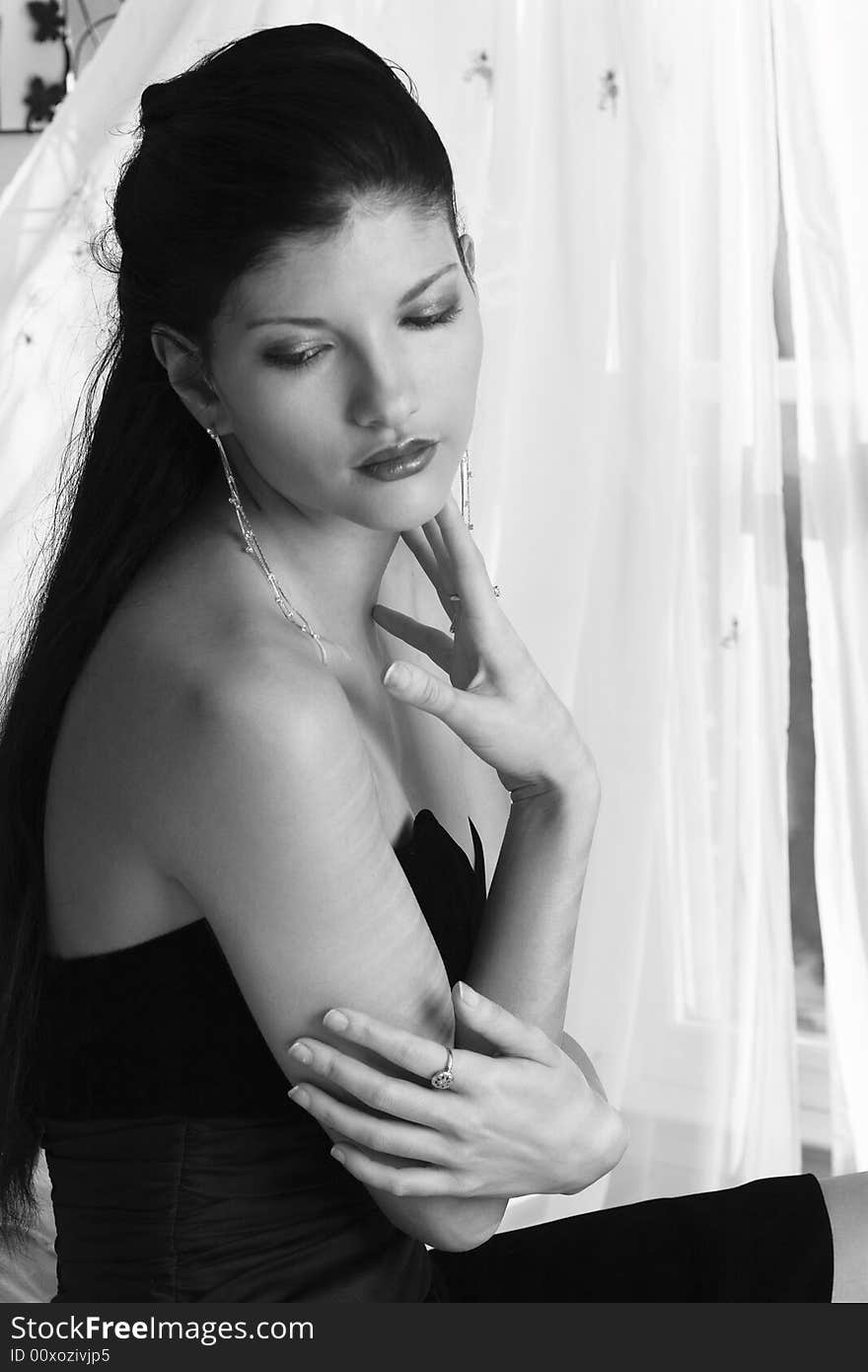 Lovely young woman wearing a black strapless dress sitting in front of a window with lace curtains. Lovely young woman wearing a black strapless dress sitting in front of a window with lace curtains