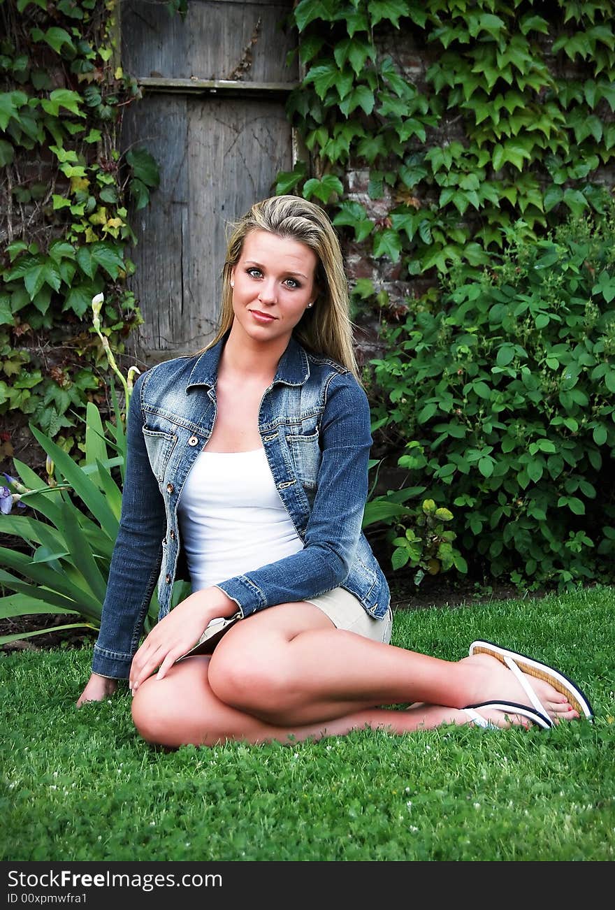 Attractive blond woman in a blue denim jacket sitting down on a lush green lawn. Vertically framed shot. Attractive blond woman in a blue denim jacket sitting down on a lush green lawn. Vertically framed shot.