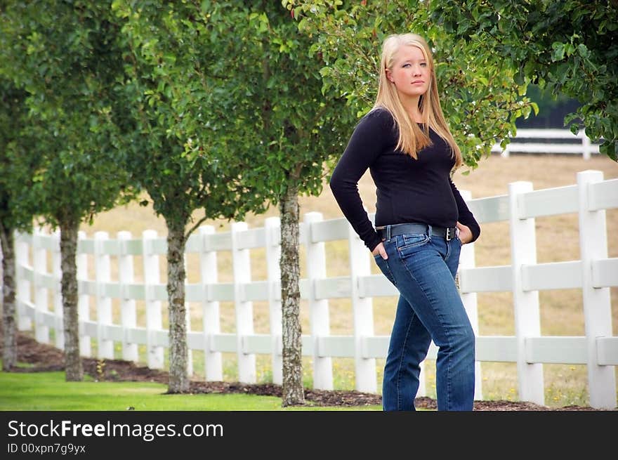 Outdoor shot of a teenage girl, with blonde hair and blue eyes, standing next to white fence and green trees. Outdoor shot of a teenage girl, with blonde hair and blue eyes, standing next to white fence and green trees.