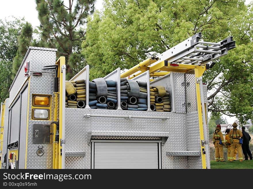 A detailed look at the back of a fire truck with hoses, ladders & grab-bars.  Fire Fighers are standing off to the right side overlooking a canyon where there is potential for wildfire. A detailed look at the back of a fire truck with hoses, ladders & grab-bars.  Fire Fighers are standing off to the right side overlooking a canyon where there is potential for wildfire.