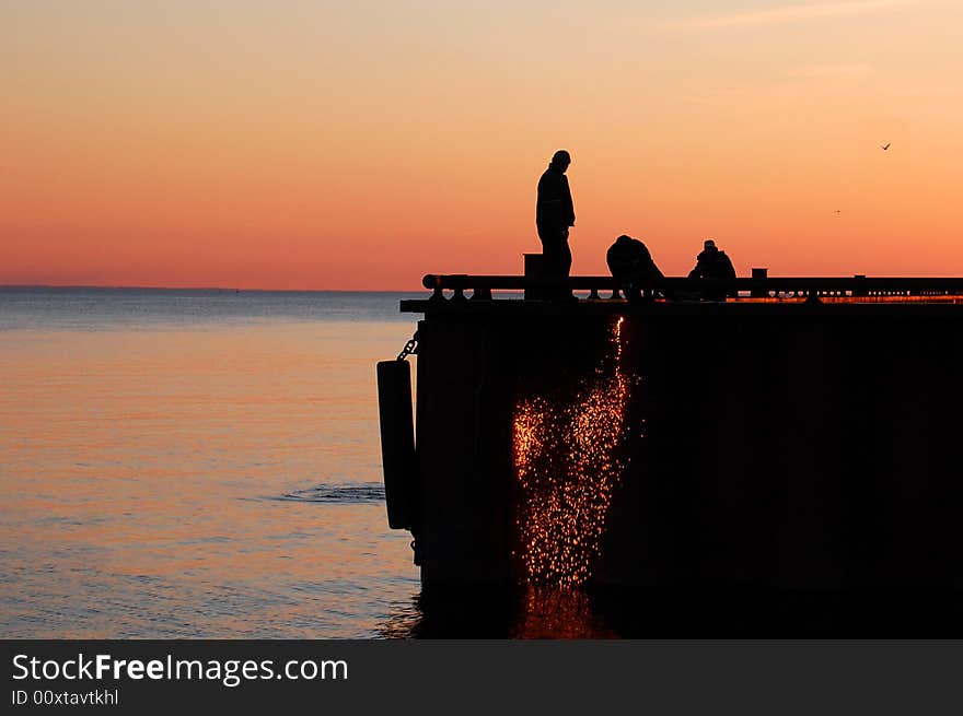 Silhouette of three workers in a port fixing a moorage, at red light. A flow of sparks is falling to the water. Silhouette of three workers in a port fixing a moorage, at red light. A flow of sparks is falling to the water.