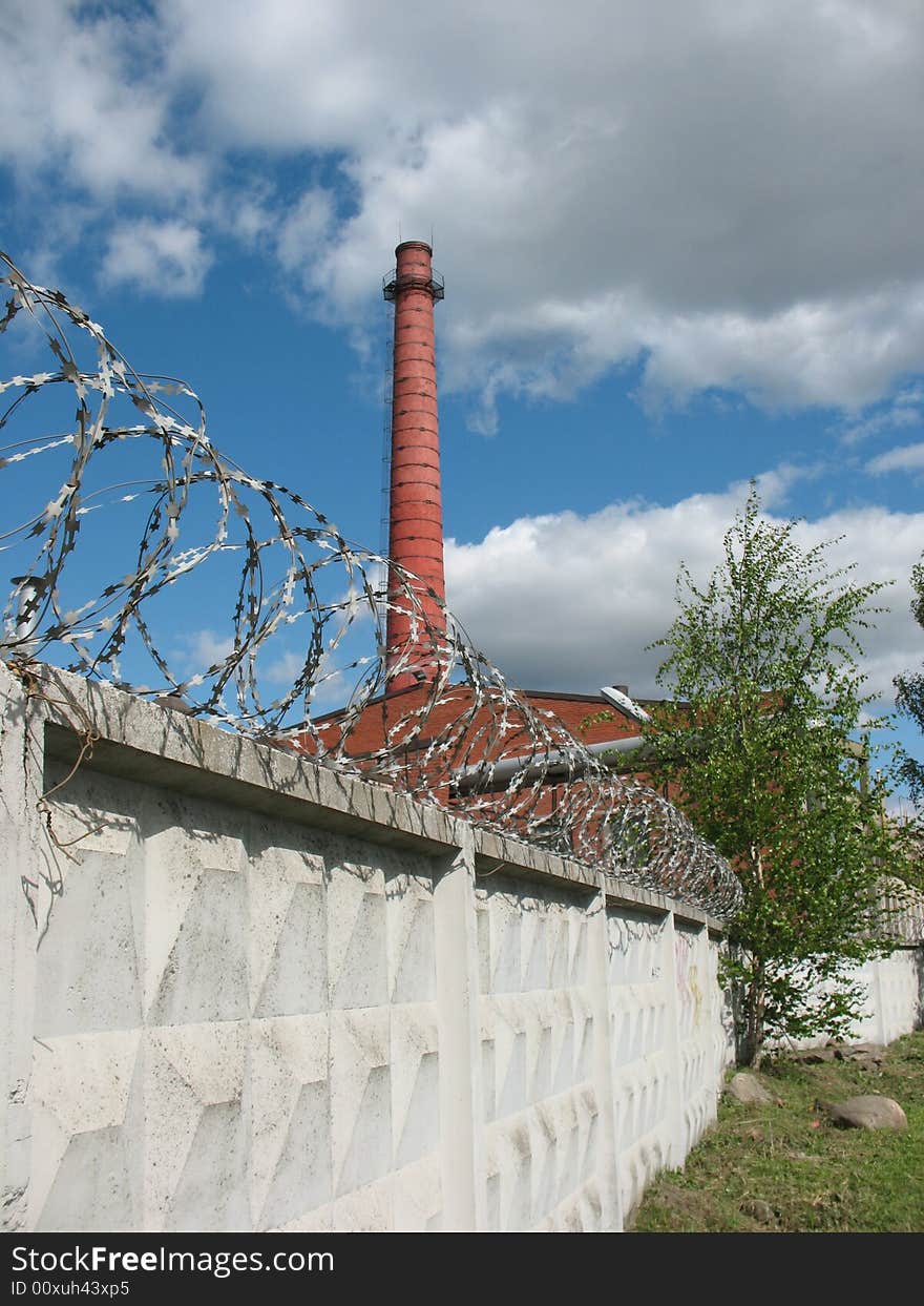 A secured industrial zone with concrete fence, barbed wire and brick chimney-stalk on the blue sky background.