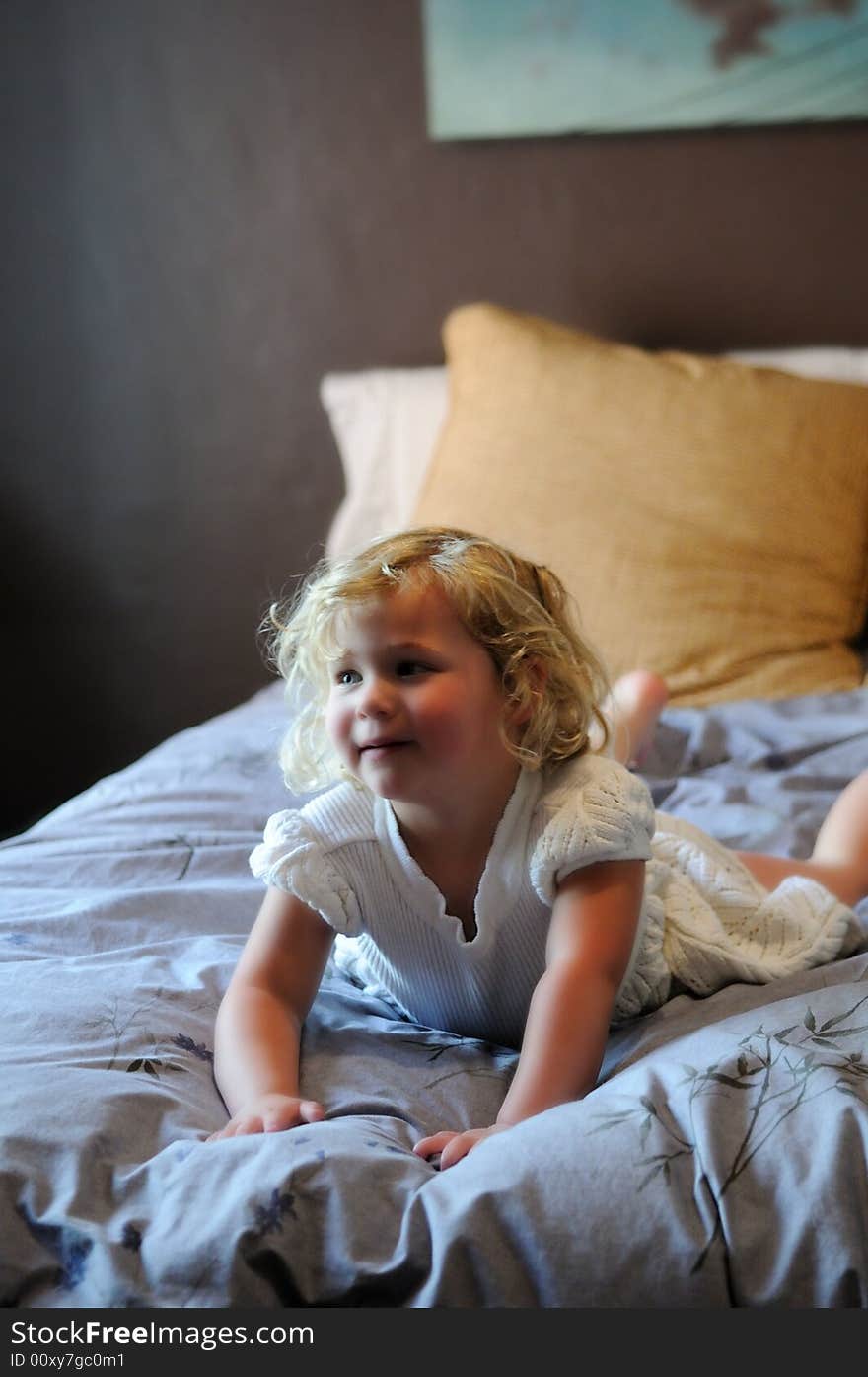 A happy little girl with blonde hair and a white dress plays on her parents bed. A happy little girl with blonde hair and a white dress plays on her parents bed.