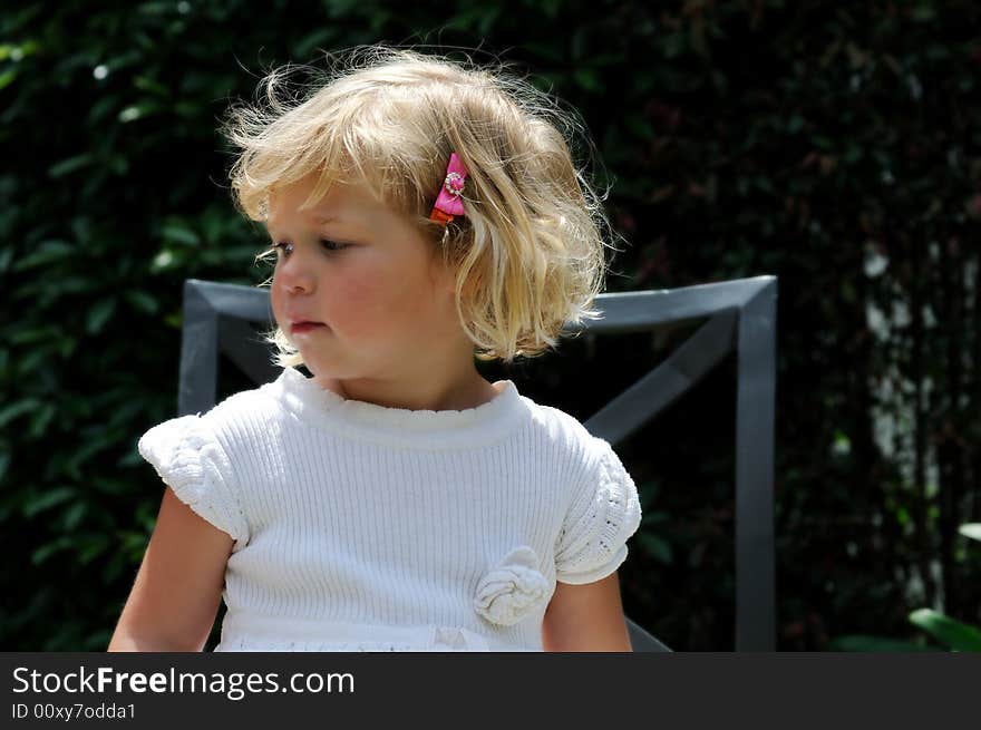 Little girl sitting outside in a garden on a chair, gazing pensively off to the side. Little girl sitting outside in a garden on a chair, gazing pensively off to the side.