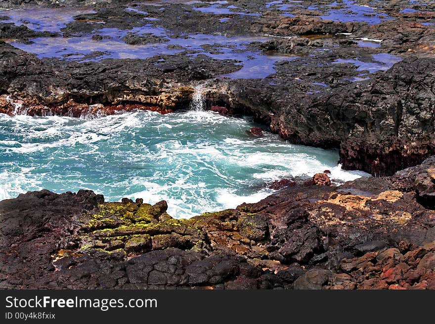 Ocean wave splashing in black lava rock on beach, blue pools of water in background, in foreground, green algae on black rock and some reddish rocks. Ocean wave splashing in black lava rock on beach, blue pools of water in background, in foreground, green algae on black rock and some reddish rocks