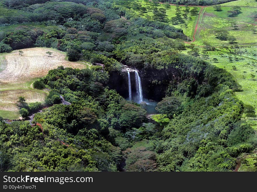 Two waterfalls in Kauai, green tropical vegetation surrounding the falls, to the right is grassy area with scattered trees, to the left is dried grassy field that has been mowed. Two waterfalls in Kauai, green tropical vegetation surrounding the falls, to the right is grassy area with scattered trees, to the left is dried grassy field that has been mowed