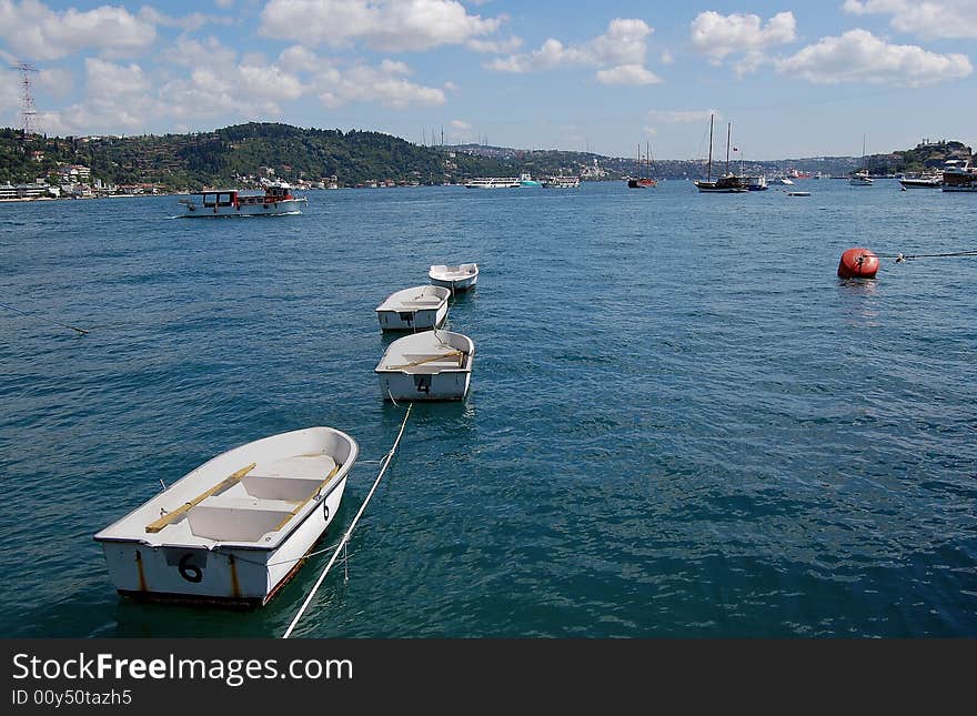 A small fleet of four numbered white dinghies of decreasing sizes tied together on blue waters of the Bosphorus. Also in view are various boats, a red float, and blue sky with partial clouds. A small fleet of four numbered white dinghies of decreasing sizes tied together on blue waters of the Bosphorus. Also in view are various boats, a red float, and blue sky with partial clouds.