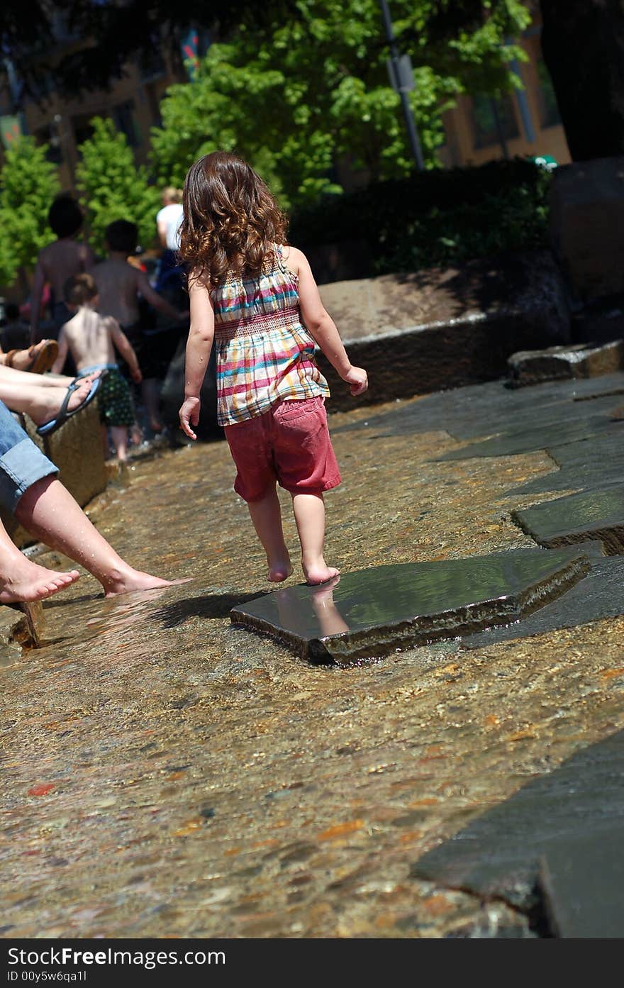 Cute young girl playing in a public fountain on a summer day. Vertically framed shot with the little girl facing away from the camera.