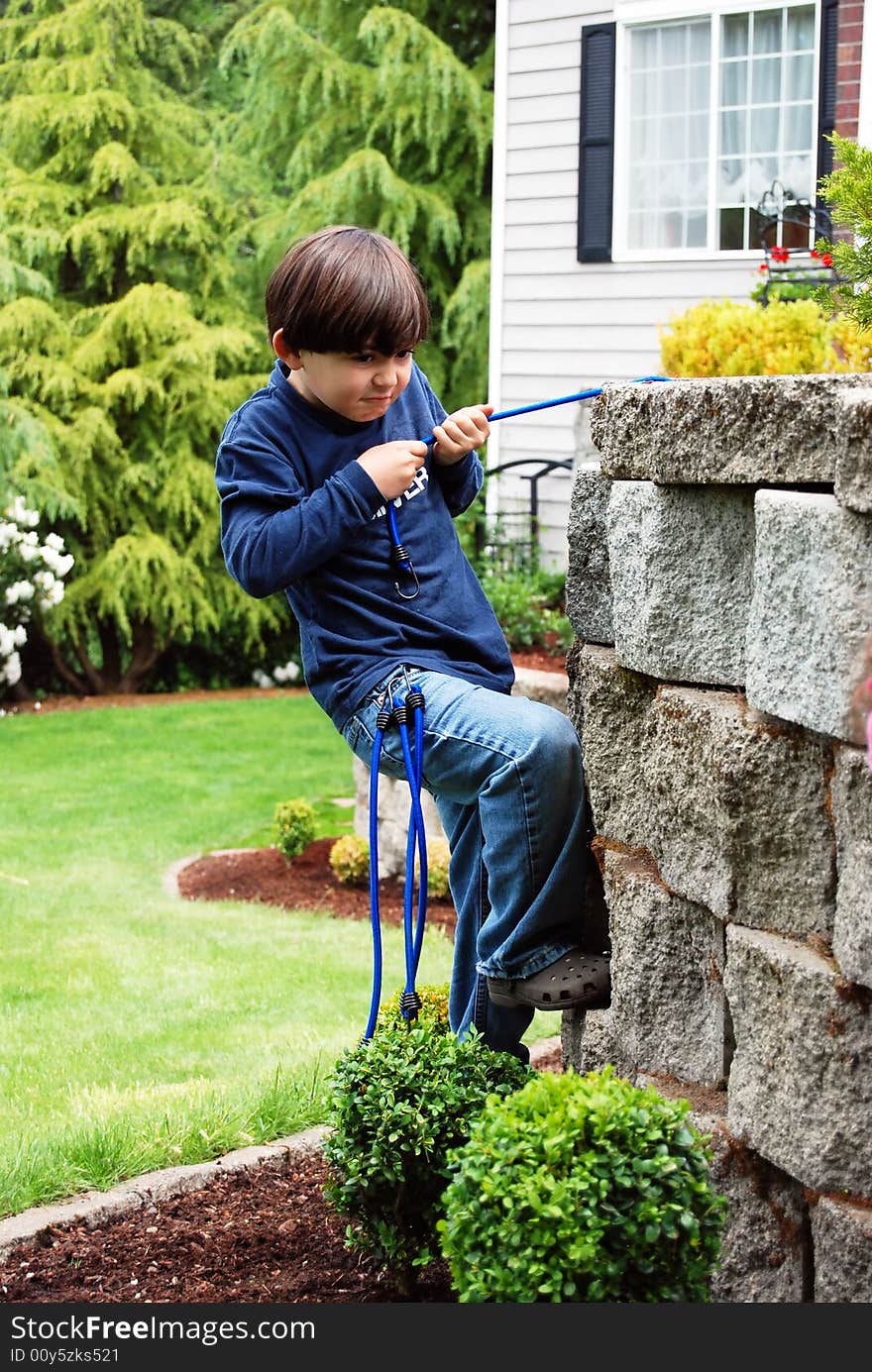 Adorable little boy top roping and climbing a stone garden wall using bungee cords as climbing rope. Vertically framed shot in which he looks focused his goal. Adorable little boy top roping and climbing a stone garden wall using bungee cords as climbing rope. Vertically framed shot in which he looks focused his goal.