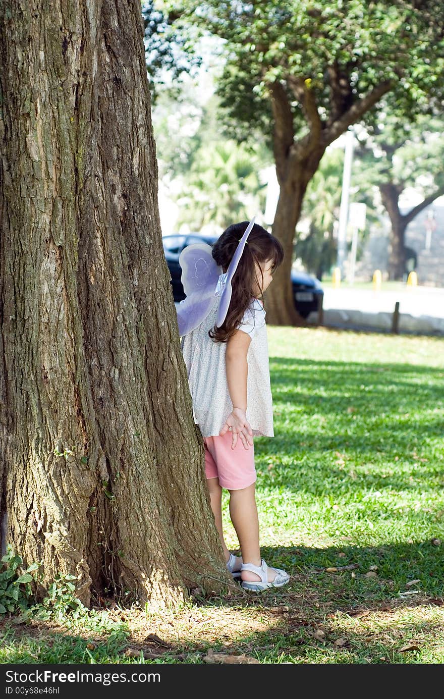 Cute little girl wearing butterfly wings leaning against a tree in sunny green park. Vertically framed outdoor shot with the girl facing away from the camera. Cute little girl wearing butterfly wings leaning against a tree in sunny green park. Vertically framed outdoor shot with the girl facing away from the camera.