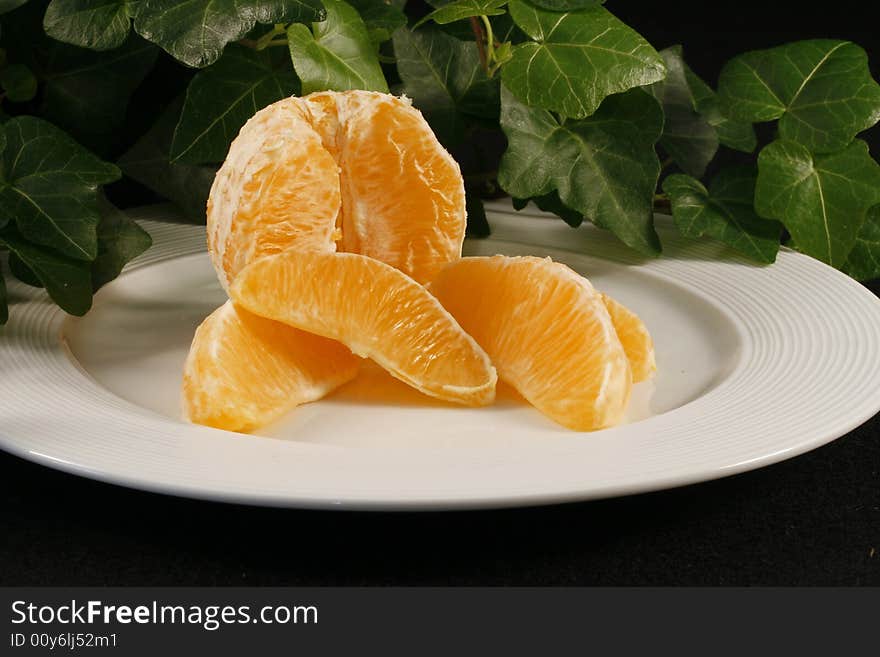 A display of a peeled orange with orange wedges on a white plate which is sitting on a black cloth.  There is ivy in the background.