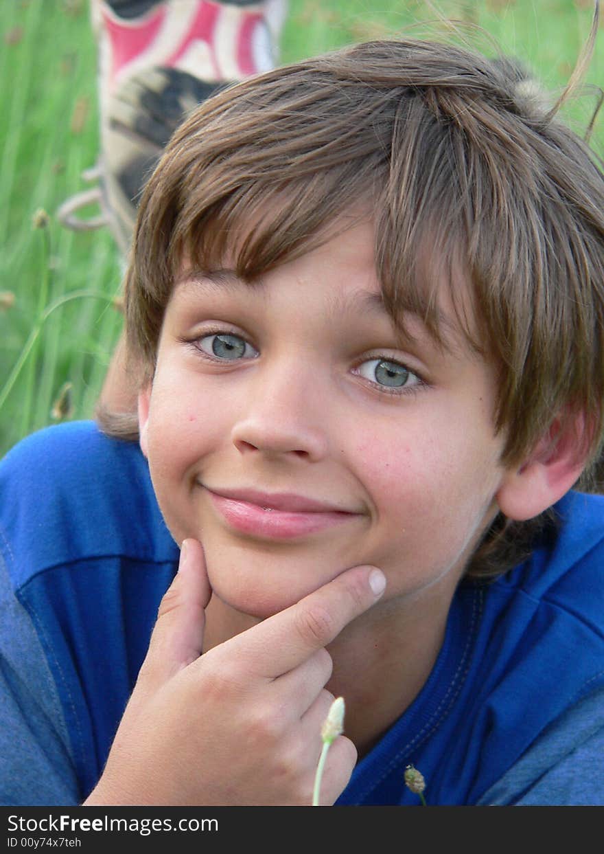 Boy in blue t-shirt with fingers on chin; he is lying in the green grass/field. Boy in blue t-shirt with fingers on chin; he is lying in the green grass/field