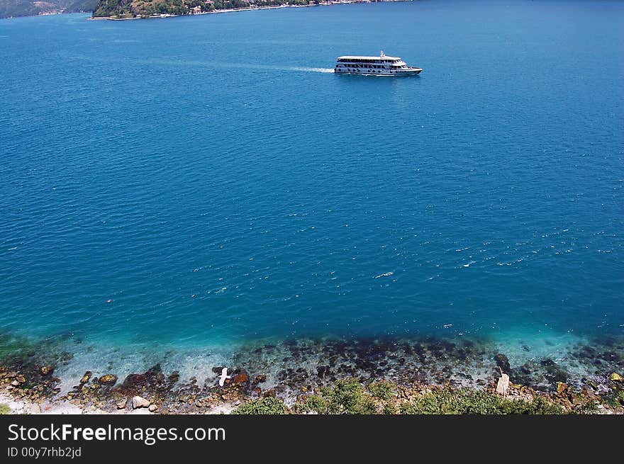 Bird-eye view of a lonely boat cruising on the blue sea. The shoreline, a seagull, and the coastal residences are also in view. Bird-eye view of a lonely boat cruising on the blue sea. The shoreline, a seagull, and the coastal residences are also in view.