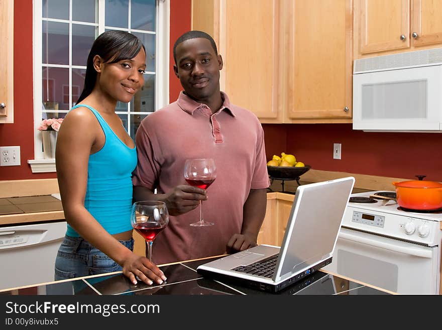 Attractive African American couple, with neutral expressions, standing in a kitchen, holding wine glasses, while using a laptop. Horizontally framed shot with the man and woman looking at the camera. Attractive African American couple, with neutral expressions, standing in a kitchen, holding wine glasses, while using a laptop. Horizontally framed shot with the man and woman looking at the camera.