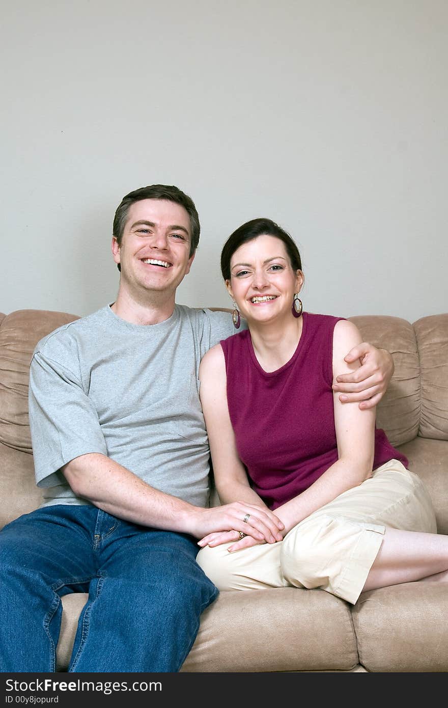 Attractive couple sitting on their living room couch with their arms around each other. Both are smiling at the camera. Vertically framed shot. Attractive couple sitting on their living room couch with their arms around each other. Both are smiling at the camera. Vertically framed shot.