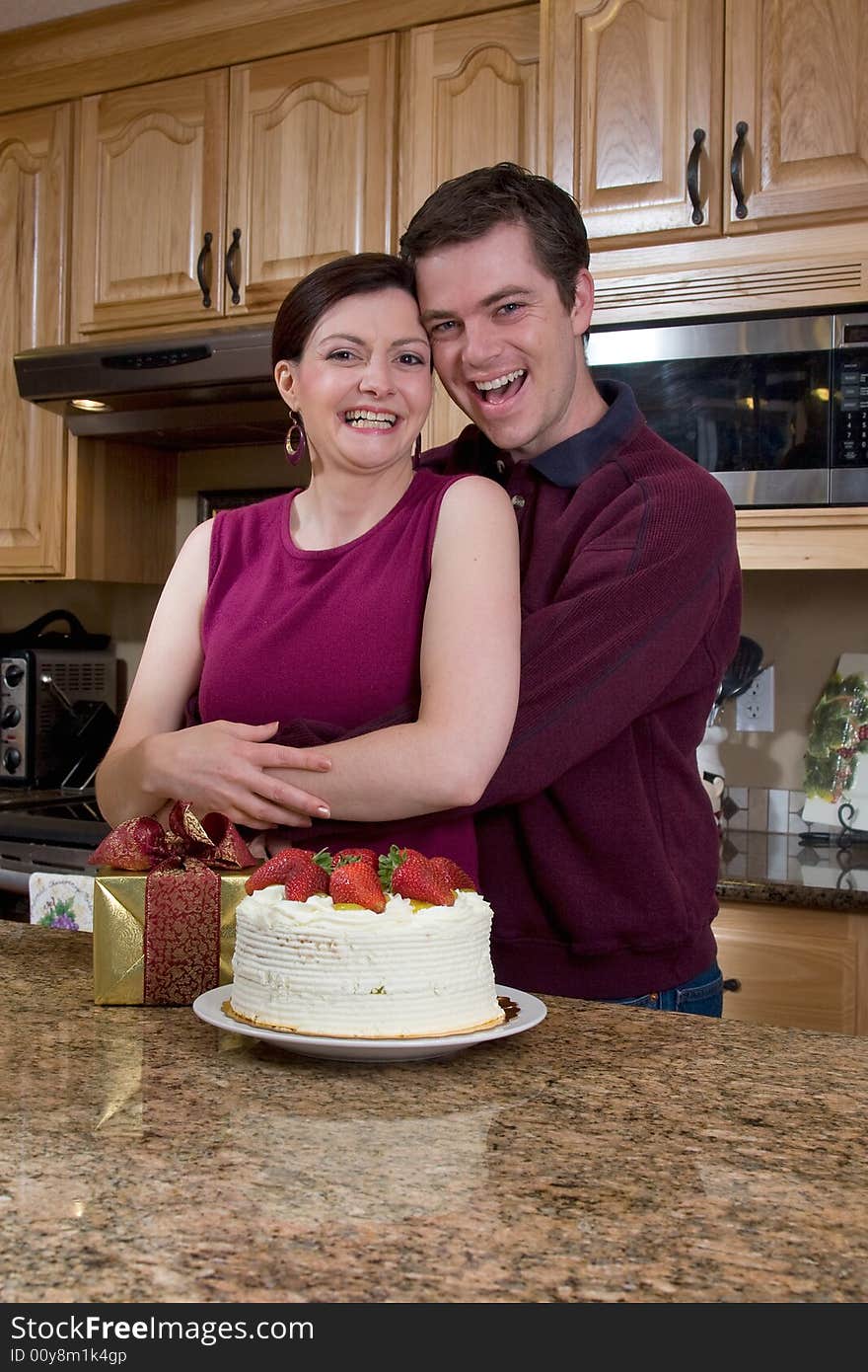 Attractive couple hugging in the kitchen by a counter covered with a cake and a present. Pair are laughing heartily and looking at the camera. Vertically framed shot. Attractive couple hugging in the kitchen by a counter covered with a cake and a present. Pair are laughing heartily and looking at the camera. Vertically framed shot.