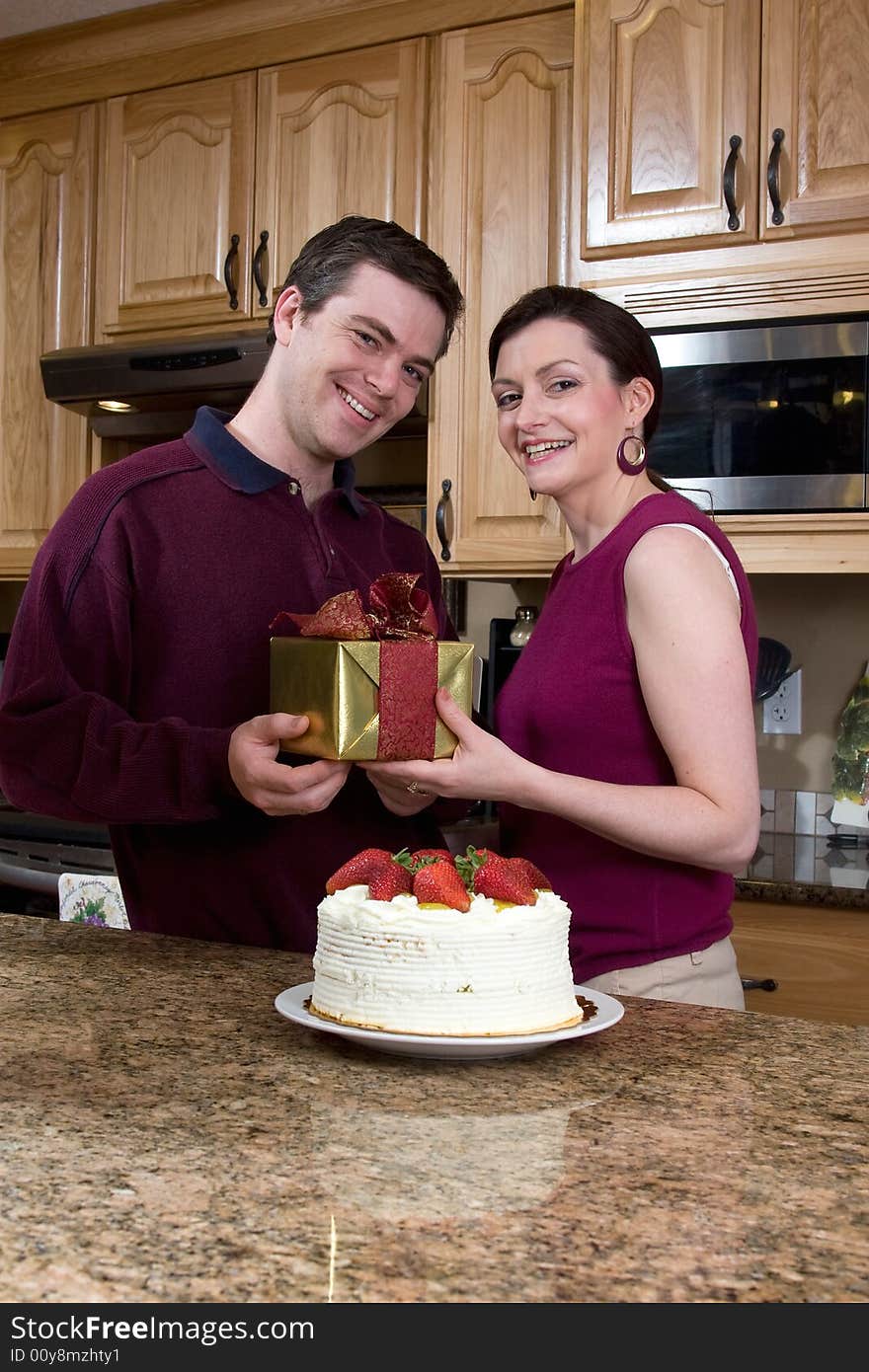 Attractive couple laughing at the camera while standing in the kitchen and exchanging gifts and cake. Vertically framed shot. Attractive couple laughing at the camera while standing in the kitchen and exchanging gifts and cake. Vertically framed shot.