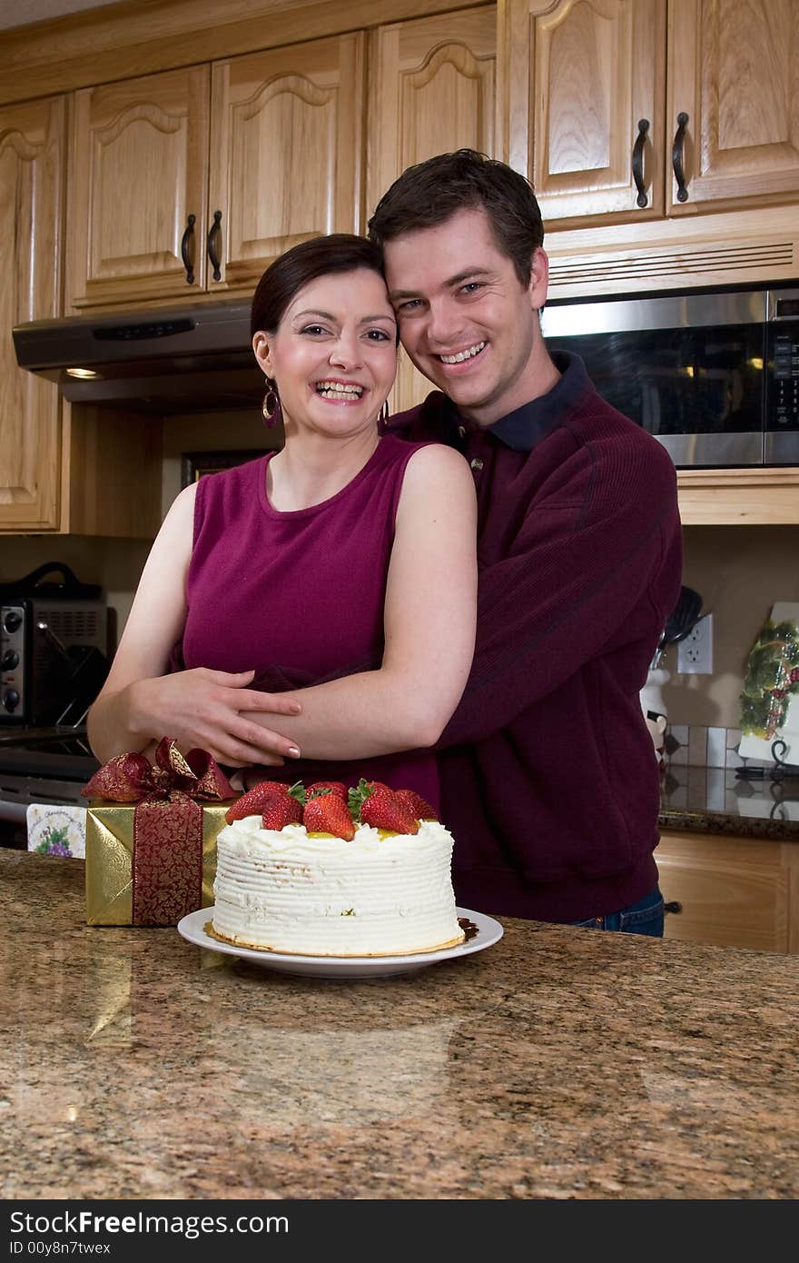 Attractive couple hugging in the kitchen by a counter covered with a cake and a present. Vertically framed shot. Pair are smiling at the camera. Attractive couple hugging in the kitchen by a counter covered with a cake and a present. Vertically framed shot. Pair are smiling at the camera.