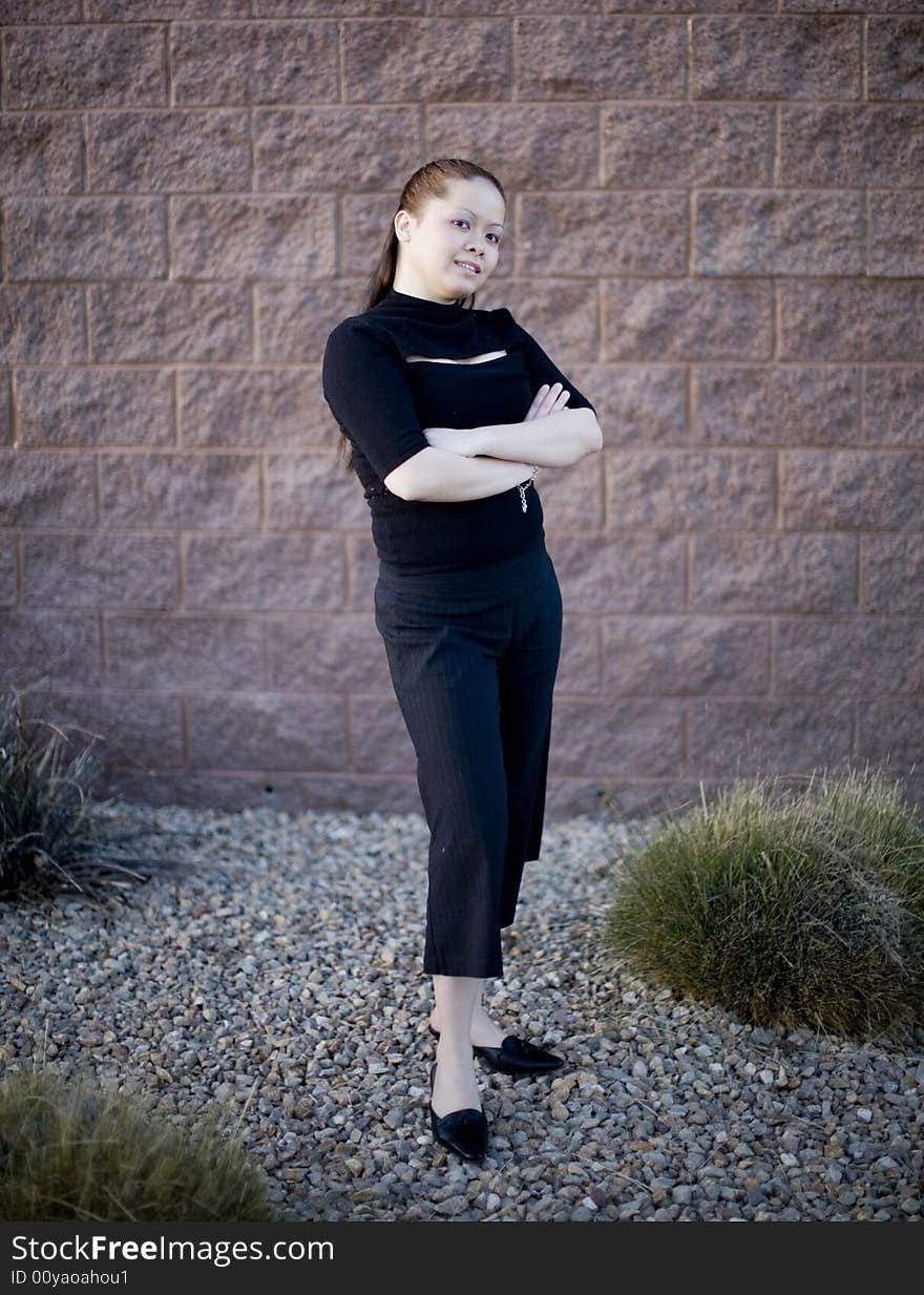 Young Asian Woman standing outside against a brick wall with her arms crossed while wearing a black business outfit