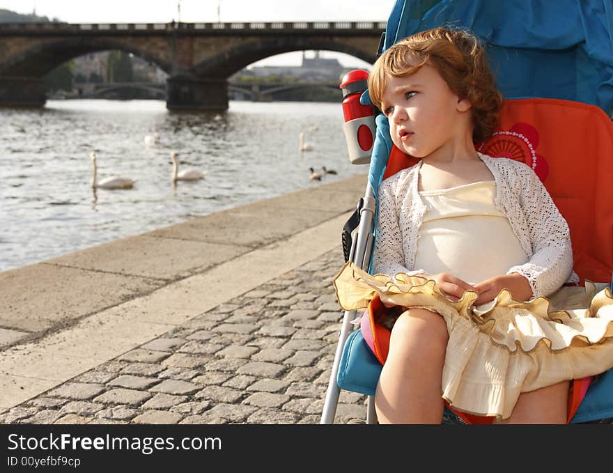 A little girl is sitting in a carriage on a river embankment and watching the swans.