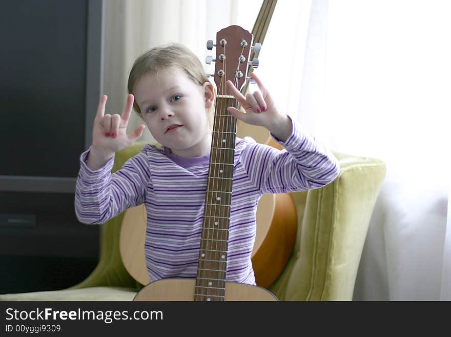 Female child age six holding an acoustic guitar and gesturing the rock on sign with her hands. The child is wearing a purple striped shirt and sitting on a green chair. Female child age six holding an acoustic guitar and gesturing the rock on sign with her hands. The child is wearing a purple striped shirt and sitting on a green chair.