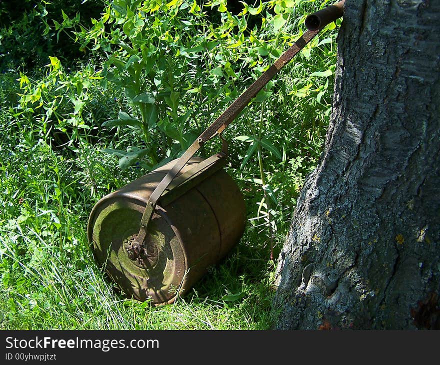 A garden lawn roller propped against a tree trunk cast in dappled sunlight.